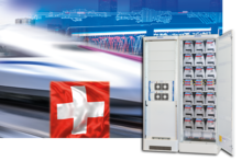 In the background an assembly with a train and a national flag of Switzerland. In the front an ENERTRONIC modular SE with 20 kVA modules and a modular battery cabinet.